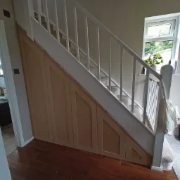 Drewniane szafki pod schodami w przedpokoju, z białymi schodami i poręczą, widok na okno z rośliną doniczkową na parapecie, oraz torba podróżna i kapcie na dywaniku.