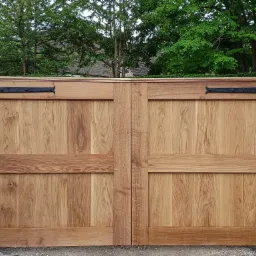 Rustykalne dwuskrzydłowe drzwi drewniane z czarnymi okuciami, osadzone w kamiennych słupkach, na tle zieleni drzew.