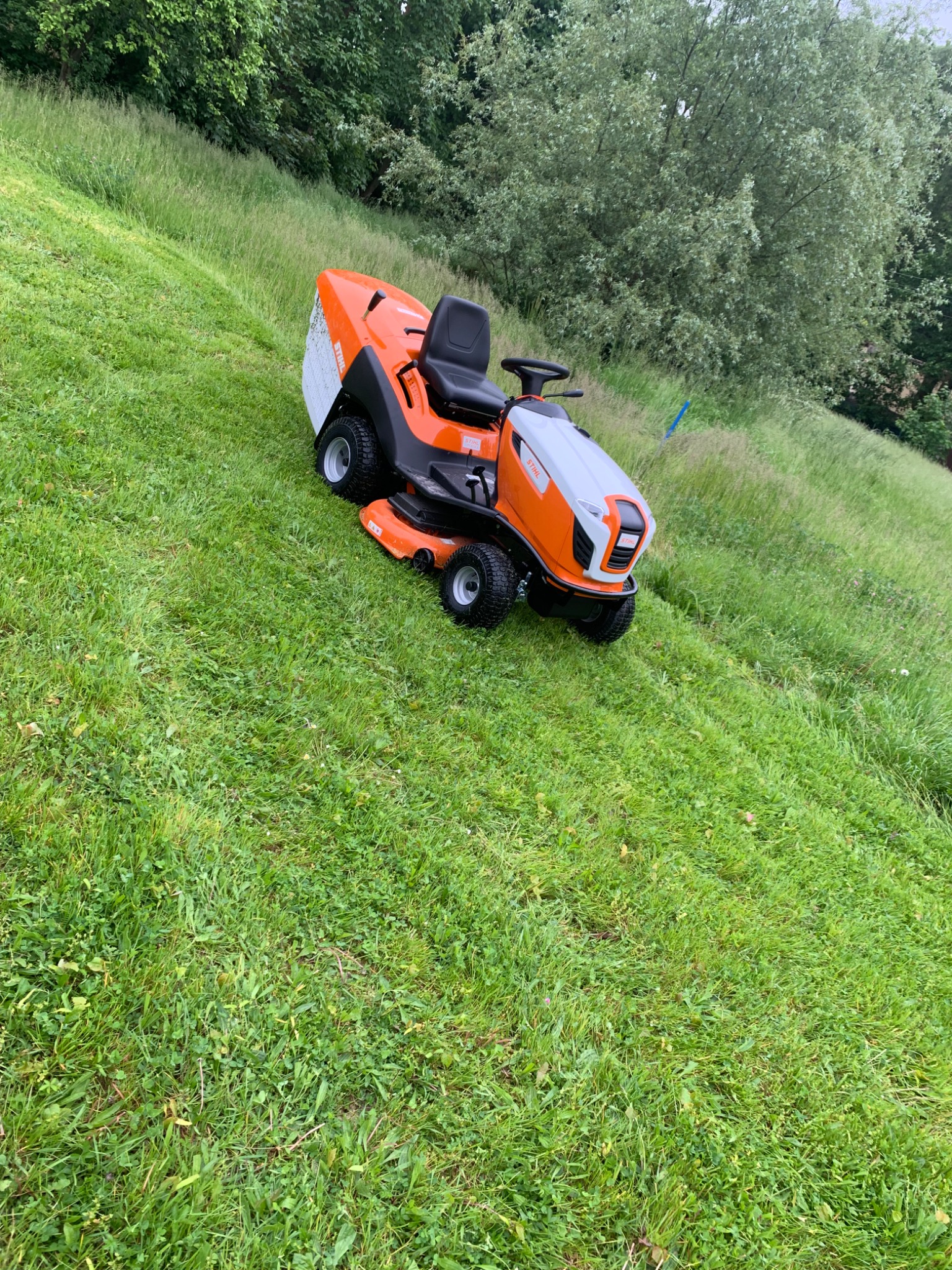 Pomarańczowy traktorek do koszenia trawy Stihl na zielonym, nierównym trawniku z wysoką trawą w tle.