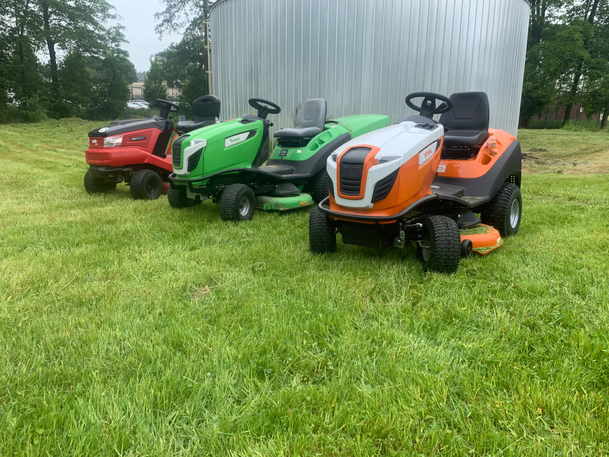 Cztery różne traktorki do trawy: czerwony Solo, dwa zielone Viking i pomarańczowo-biały Stihl, stoją zaparkowane na trawniku przed srebrnym silosie.