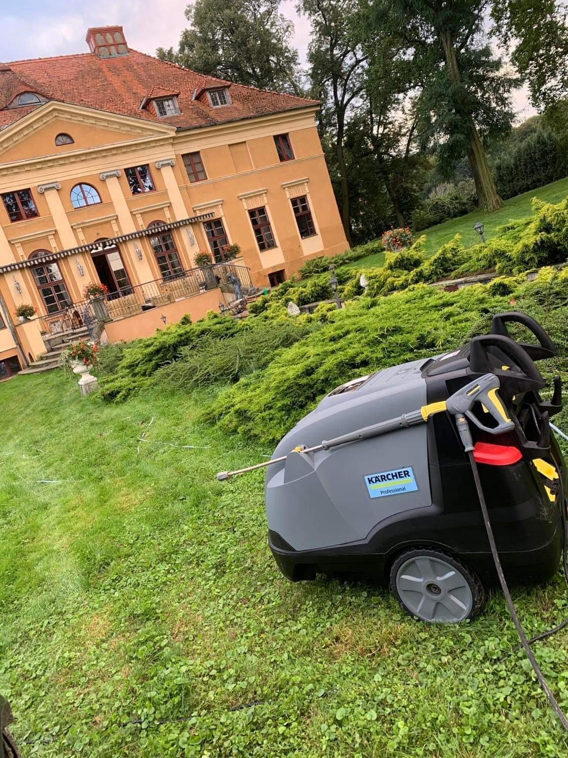 Profesjonalna myjka ciśnieniowa Karcher na trawniku przed okazałą, dwupiętrową willą z elementami dekoracyjnymi i zadbanym ogrodem.