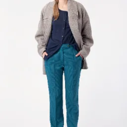 Kobieta w granatowej czapce, szarym płaszczu i turkusowych spodniach o drobnym wzorze, stojąca na białym tle z rękami w kieszeniach.