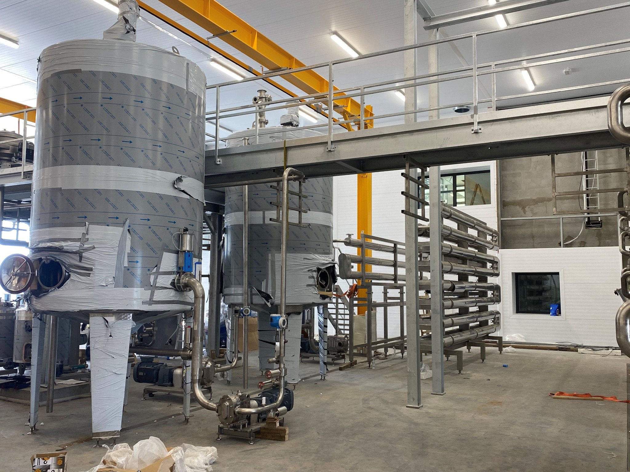 Wnętrze hali produkcyjnej z dużymi, metalowymi zbiornikami owiniętymi folią ochronną, widoczne rurociągi i konstrukcja wsporcza pod sufitem.