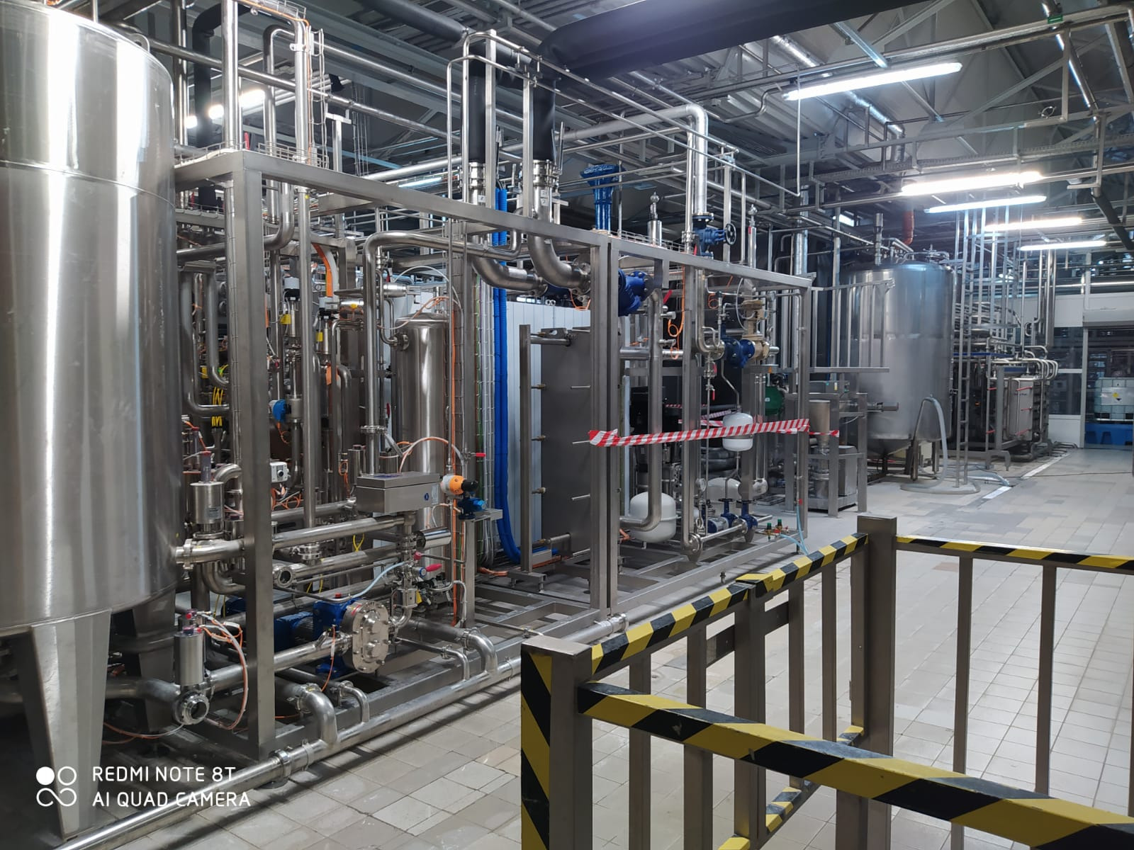 Kompleksowa instalacja przemysłowa z nierdzewnej stali z licznymi rurami, zbiornikami i zaworami, otoczona barierą ostrzegawczą z czarno-żółtymi pasami.