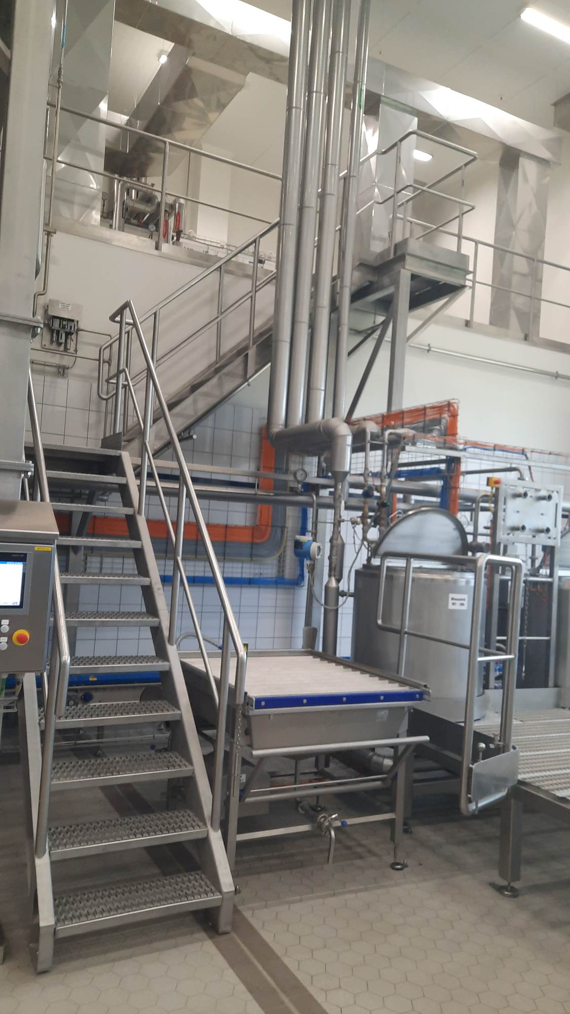 Wnętrze hali produkcyjnej z platformą roboczą ze stali nierdzewnej, schodami kratowymi i systemem rur wentylacyjnych pod sufitem.