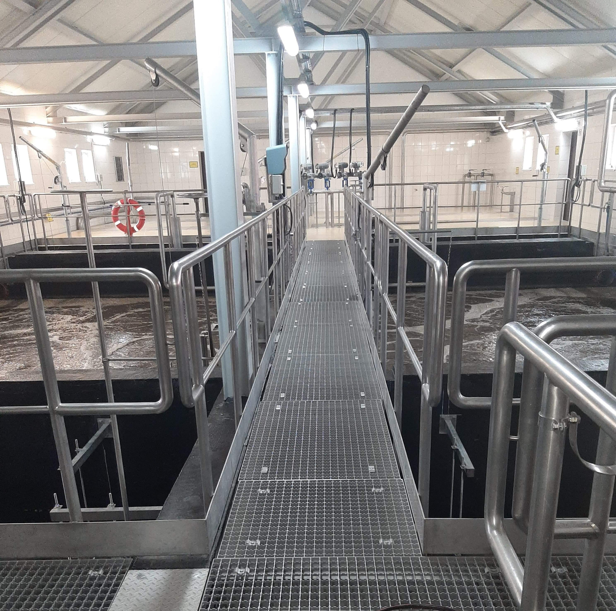 Wnętrze hali przemysłowej z metalową kładką nad zbiornikami z cieczą, widoczne rury, oświetlenie i elementy konstrukcji stalowej, w tle białe płytki i okna.