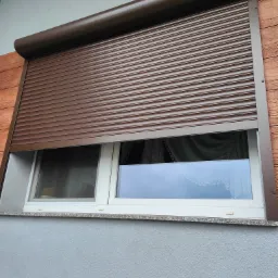 Częściowo opuszczona brązowa roleta zewnętrzna na oknie PCV w domu z elewacją w kolorze szarym i drewnianym wykończeniem.