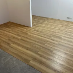 Salon z nowo ułożonymi panelami podłogowymi w odcieniach drewna, kontrastującymi z szarą, kamienną posadzką w przejściu do drugiego pomieszczenia; widoczne gniazdka elektryczne przy ścianie.