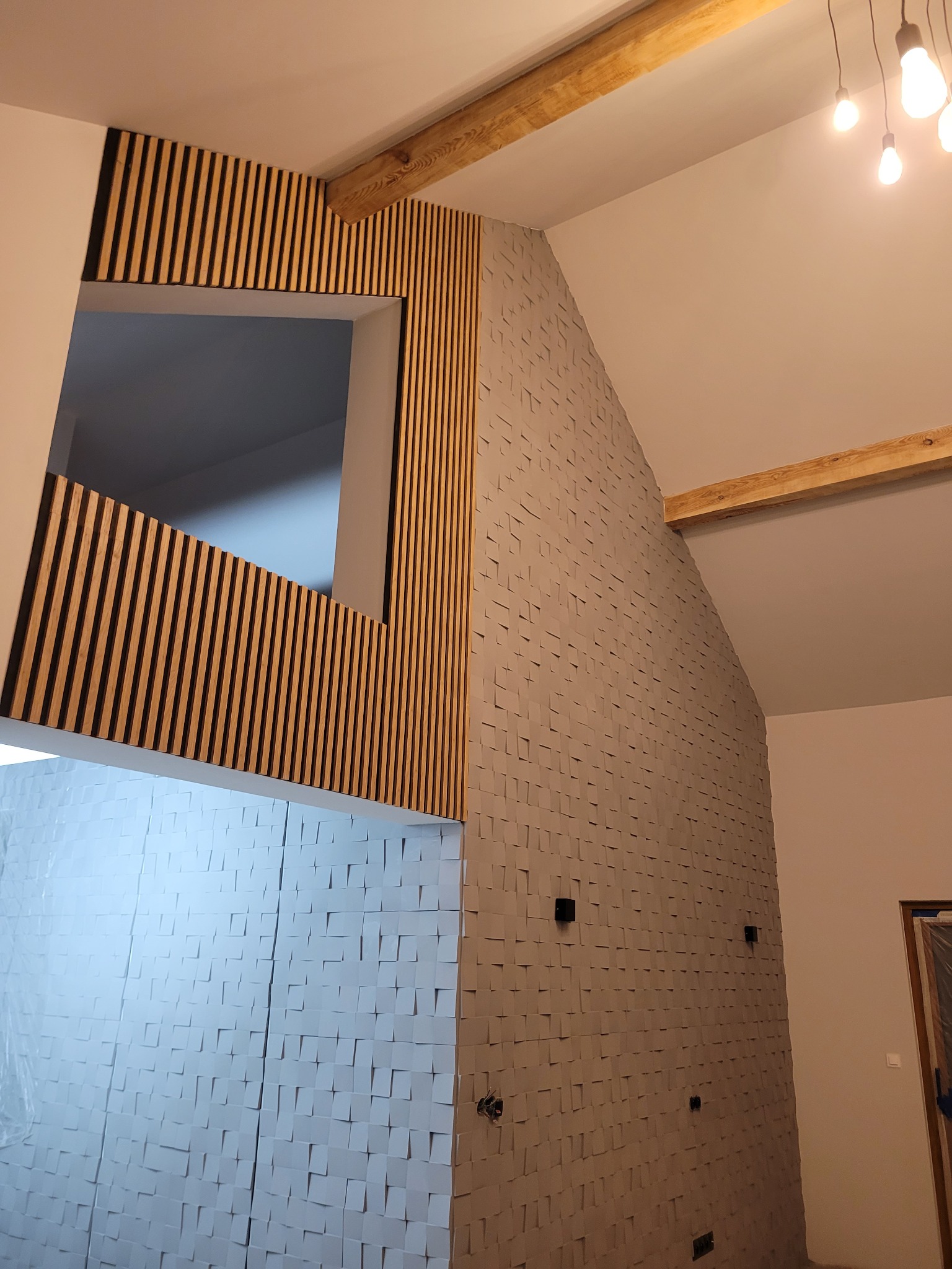 Nowoczesne wykończenie wnętrza z drewnianymi lamelami wokół okna i trójwymiarową szarą okładziną ścienną, widoczne drewniane belki stropowe i wiszące żarówki