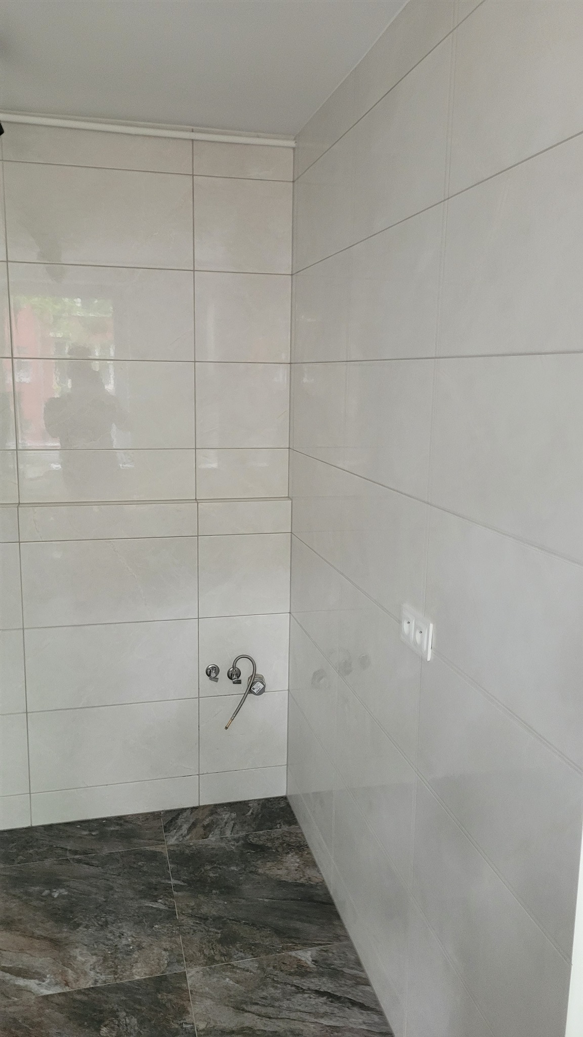 Wykończona narożna strefa łazienki z białymi, prostokątnymi płytkami ściennymi i ciemnymi, marmurkowymi płytkami podłogowymi, widoczne przyłącza wodne i gniazdko elektryczne.