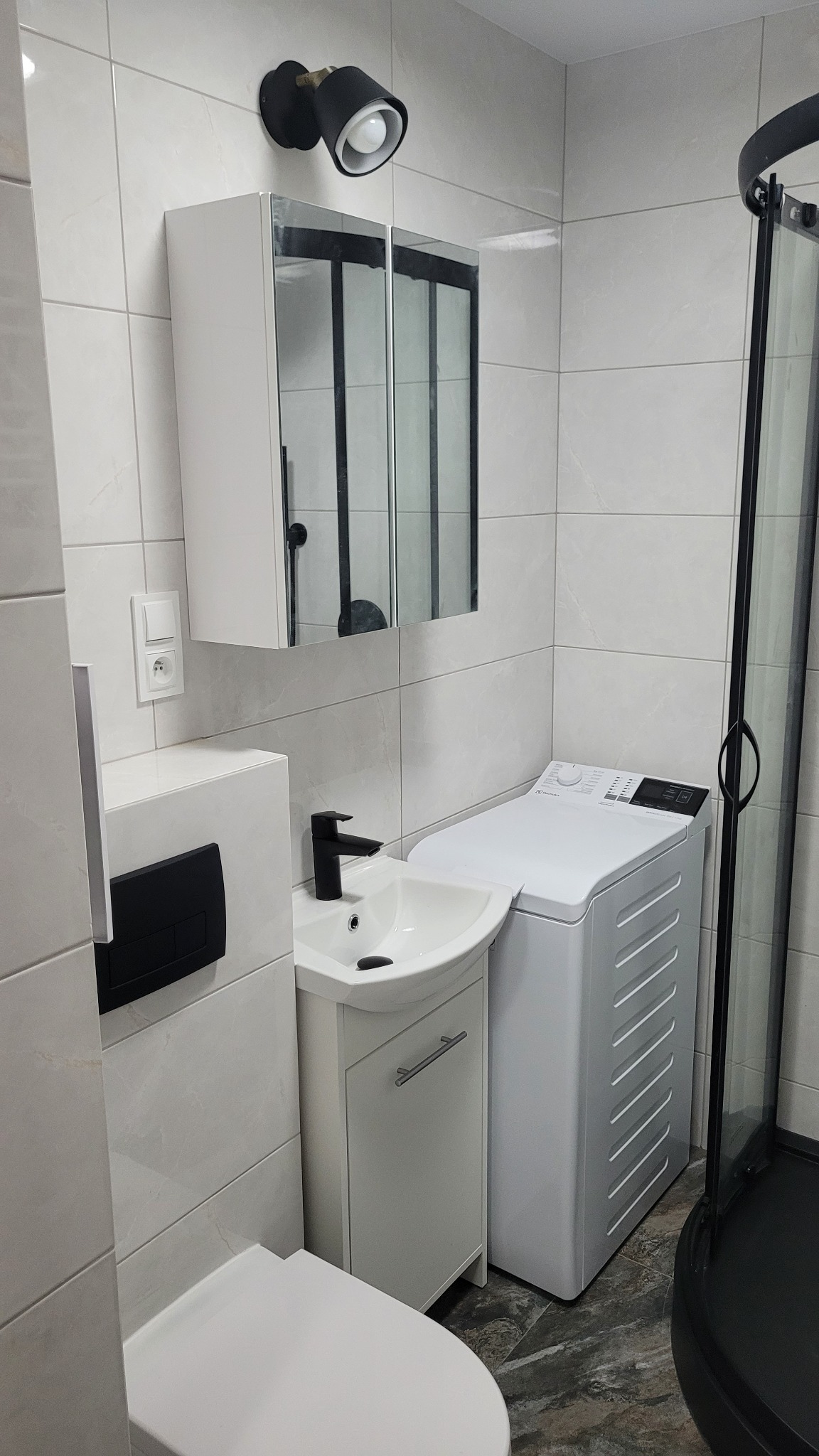Nowoczesna łazienka z umywalką na szafce, pralką i kabiną prysznicową z czarnymi elementami, oświetlona czarną lampą punktową nad szafką z lustrem, wykończona płytkami w odcieniach szarości.