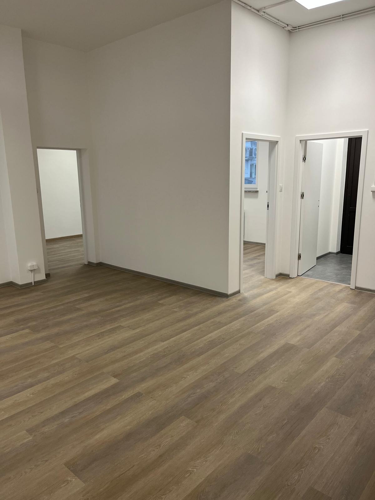 Pusty pokój z nową podłogą z paneli w odcieniach brązu, białe ściany, dwa otwarte przejścia do innych pomieszczeń z widocznymi fragmentami podłóg i ścian, minimalistyczne wykończenie wnętrza.