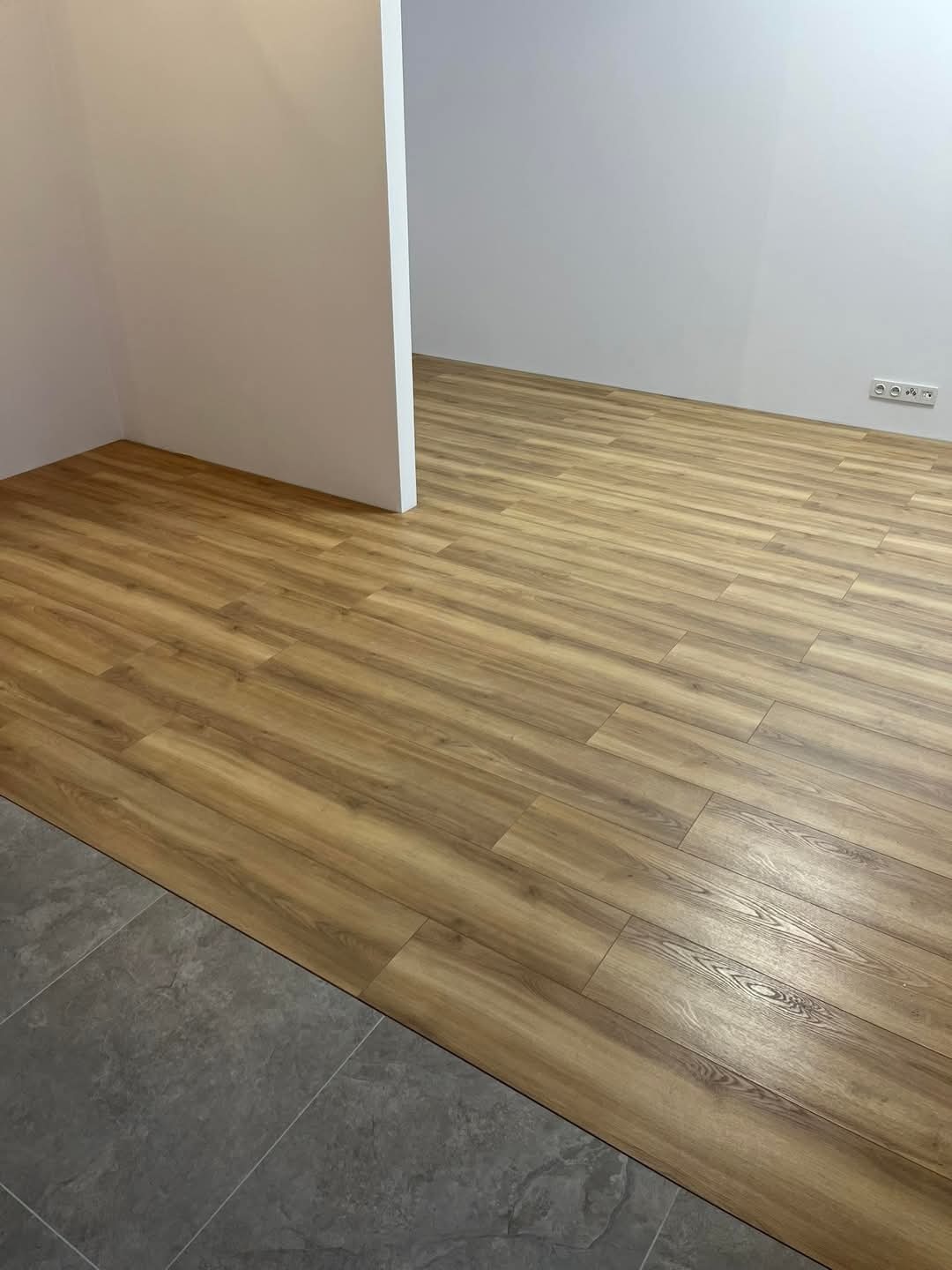 Salon z nowo ułożonymi panelami podłogowymi w odcieniach drewna, kontrastującymi z szarą, kamienną posadzką w przejściu do drugiego pomieszczenia; widoczne gniazdka elektryczne przy ścianie.