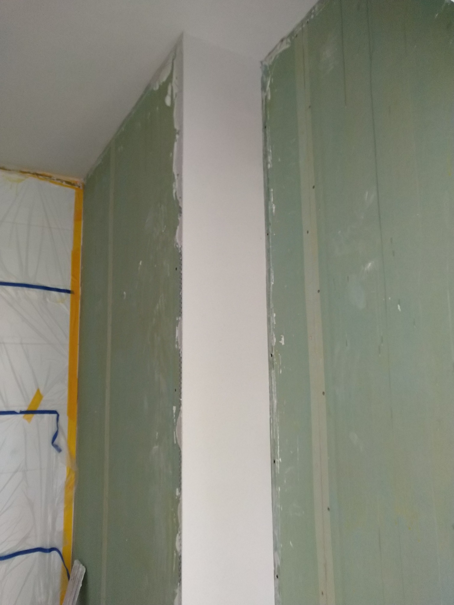 Wnętrze w trakcie prac wykończeniowych, widoczne surowe ściany z płyt gipsowo-kartonowych, częściowo pokryte białą warstwą wygładzającą, narożnik zabezpieczony listwą, folia ochronna na ścianie po...