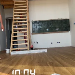 Wnętrze w trakcie prac wykończeniowych: drewniana antresola ze schodami, częściowo położona ciemnozielona glazura na ścianie, widoczne puszki elektryczne, podłoga wyłożona panelami, puszka napoju...