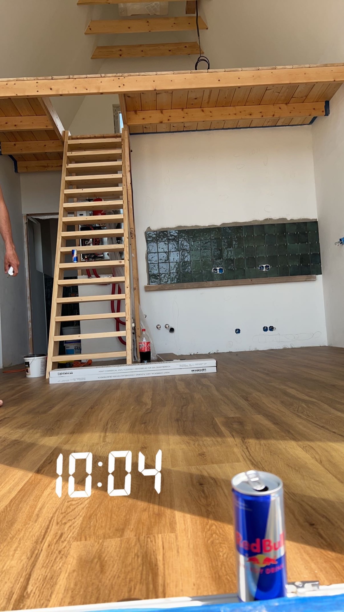 Wnętrze w trakcie prac wykończeniowych: drewniana antresola ze schodami, częściowo położona ciemnozielona glazura na ścianie, widoczne puszki elektryczne, podłoga wyłożona panelami, puszka napoju...