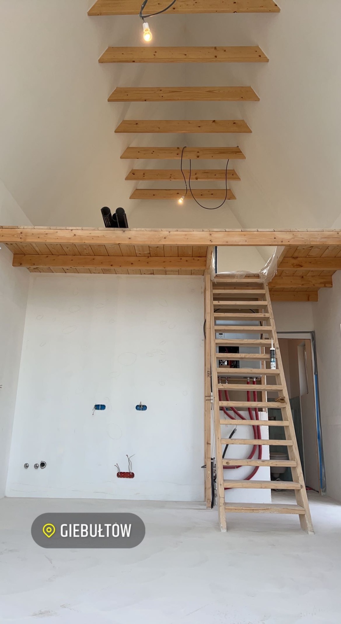 Wnętrze w trakcie wykańczania: drewniane schody prowadzą na antresolę z drewnianą balustradą, widoczne instalacje elektryczne i hydrauliczne na białych ścianach, drewniane belki pod sufitem...