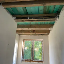 Poddasze w trakcie prac remontowych: widoczne drewniane belki stropowe, zielona podbitka z desek, przewody elektryczne przymocowane do belek, ściany w trakcie szpachlowania, okno z widokiem na zieleń.