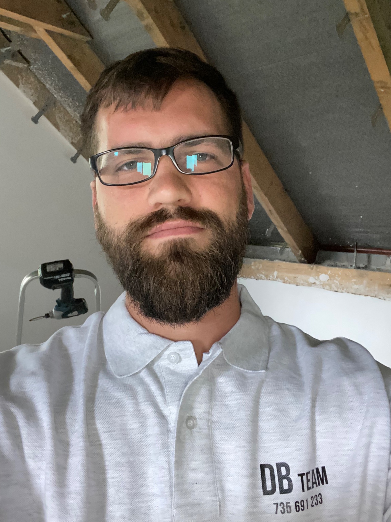 Autoportret mężczyzny w okularach i koszulce z logo firmy budowlanej na tle poddasza w trakcie prac, widoczna drabina i wkrętarka.