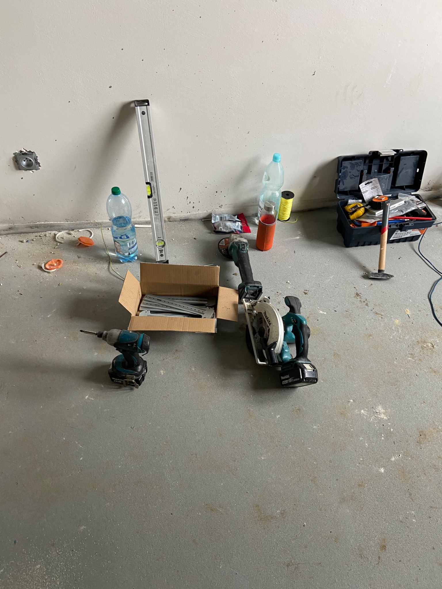 Rozłożone narzędzia budowlane na podłodze: wiertarko-wkrętarka, pilarka tarczowa, poziomica, szpachla w pudełku, skrzynka z narzędziami, butelki z wodą, wskazujące na przygotowania do prac...