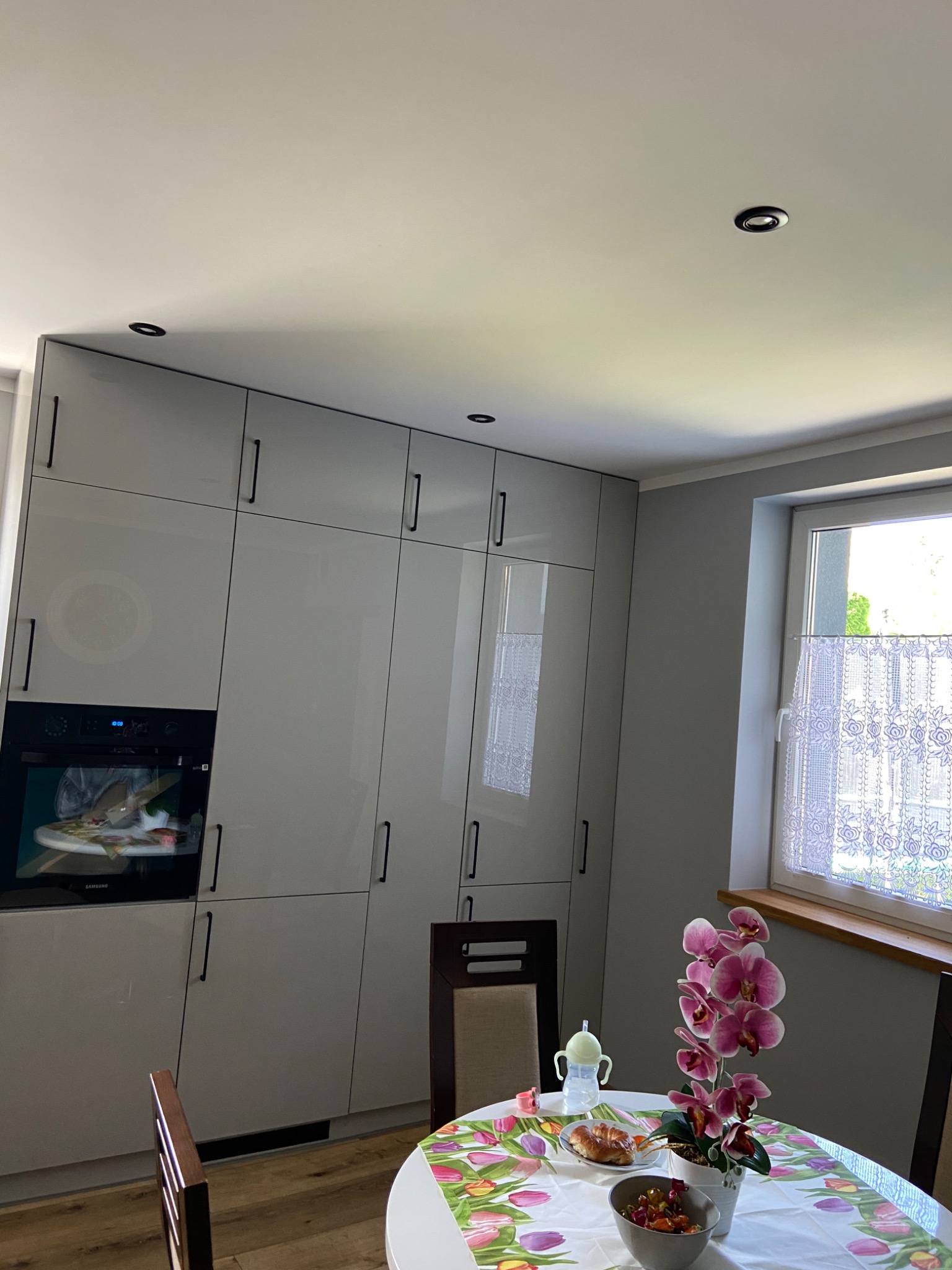 Szare, lakierowane szafki kuchenne od podłogi do sufitu z czarnymi uchwytami, wbudowany piekarnik Samsung, stół z kwiecistym obrusem, orchidea w doniczce i widok na okno z firanką.