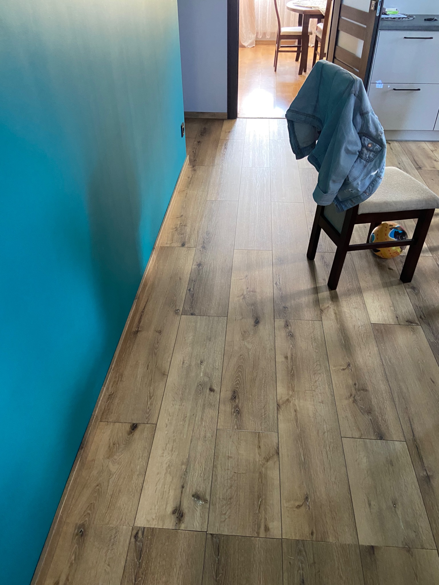 Ułożone panele podłogowe w odcieniach brązu, turkusowa ściana, krzesło z jeansową kurtką i piłką pod spodem, widok na jadalnię w tle.