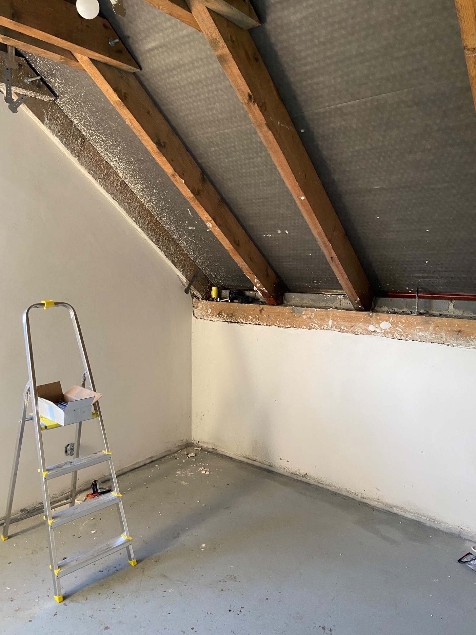 Poddasze w trakcie prac remontowych, widoczne drewniane krokwie, czarna membrana dachowa, częściowo otynkowana ściana, drabina z pudełkiem i narzędzia.