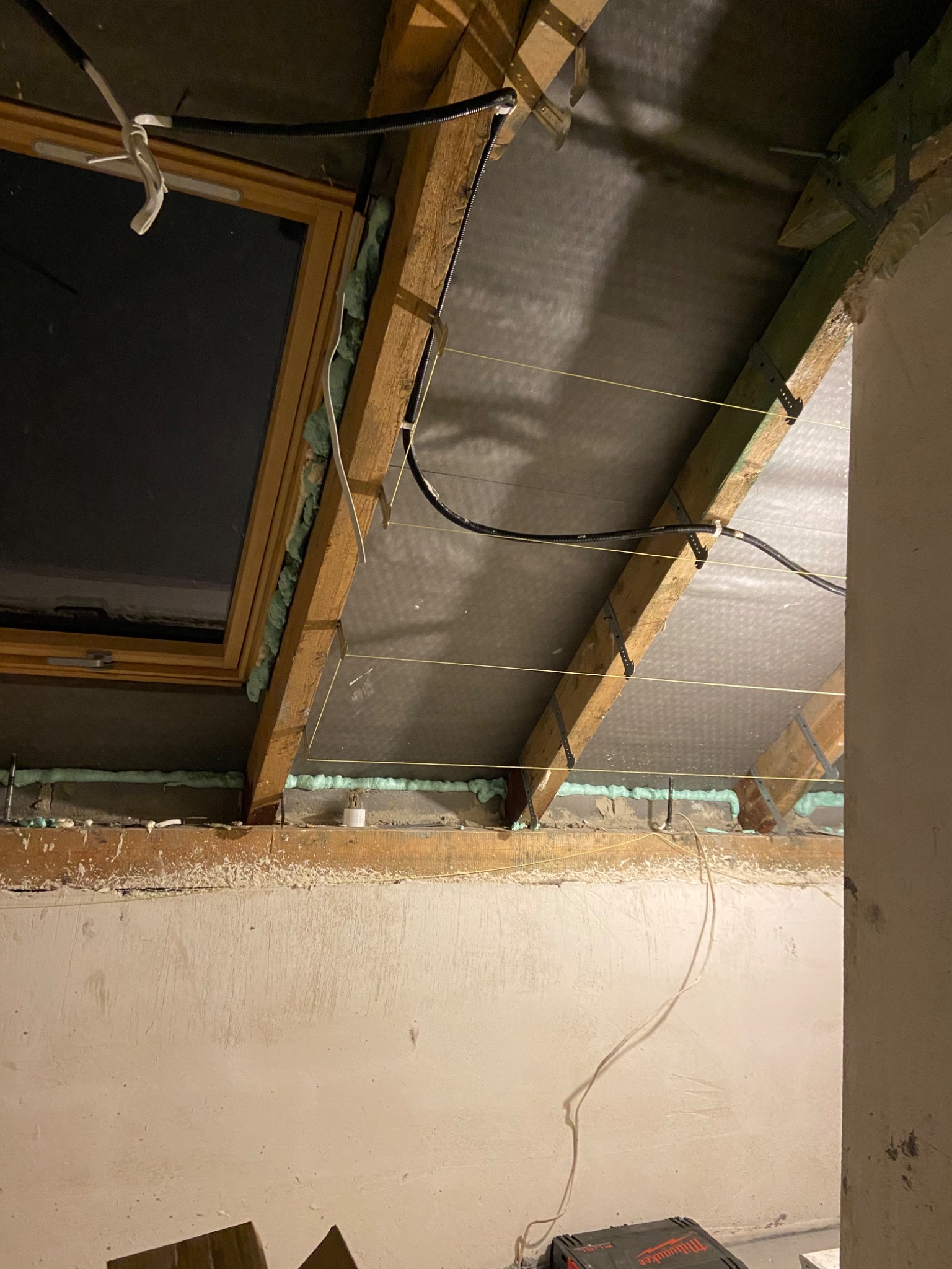 Poddasze w trakcie prac izolacyjnych: widoczne drewniane krokwie, folia paroizolacyjna, okno dachowe uszczelnione pianką montażową, oraz naprężone sznurki wyznaczające poziom.