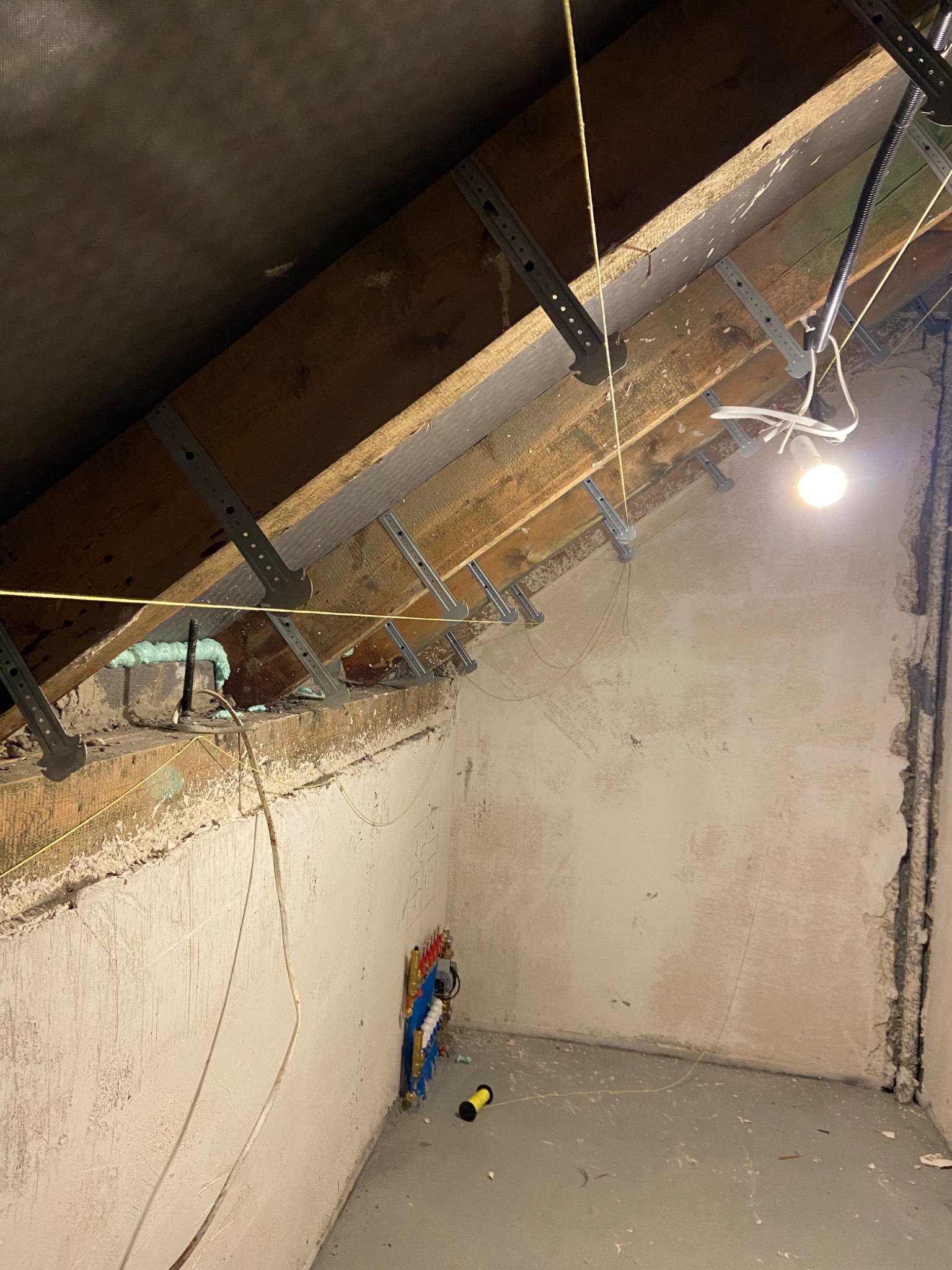 Poddasze w trakcie prac wykończeniowych: widoczne drewniane belki stropowe z zamocowanymi metalowymi uchwytami, ściany w stanie surowym z widocznymi instalacjami hydraulicznymi, oświetlenie robocze...