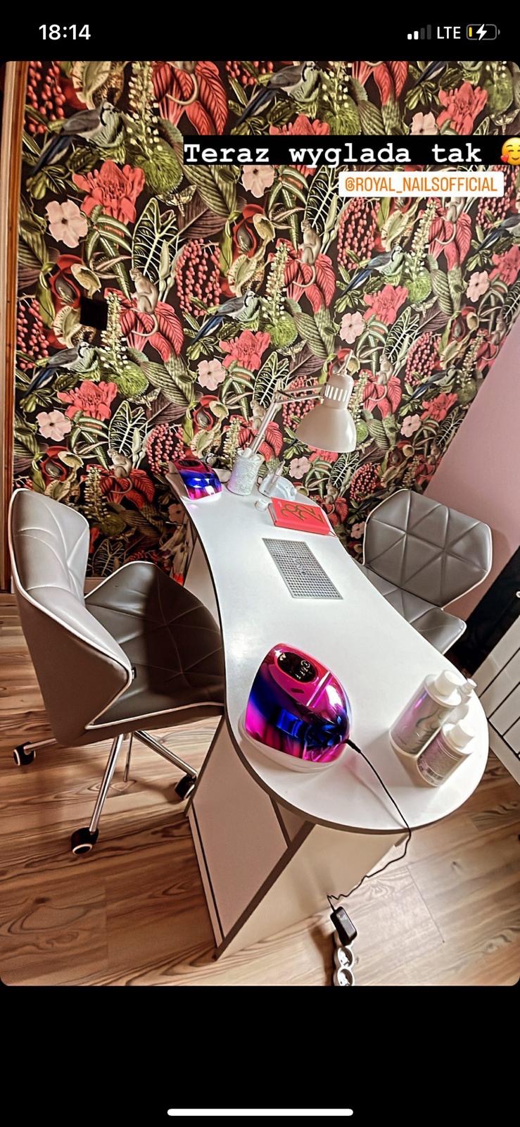 Stanowisko do manicure z lampą UV, frezarką, organizerem na lakiery, z krzesłami na tle tapety w kwiaty i ptaki.
