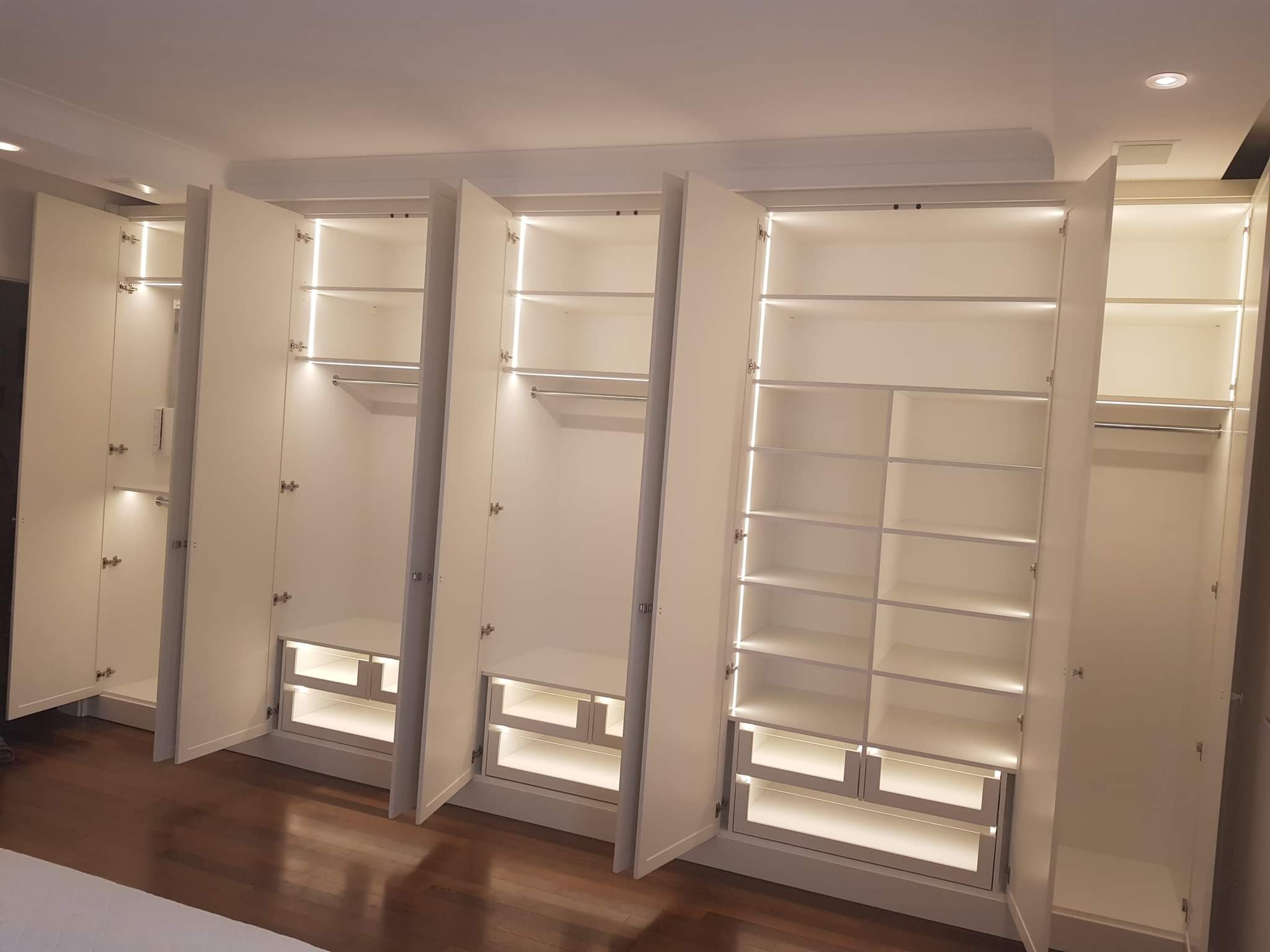 Białe, nowoczesne szafy z oświetleniem LED wewnątrz, częściowo otwarte drzwi, widoczny drewniany parkiet na podłodze.
