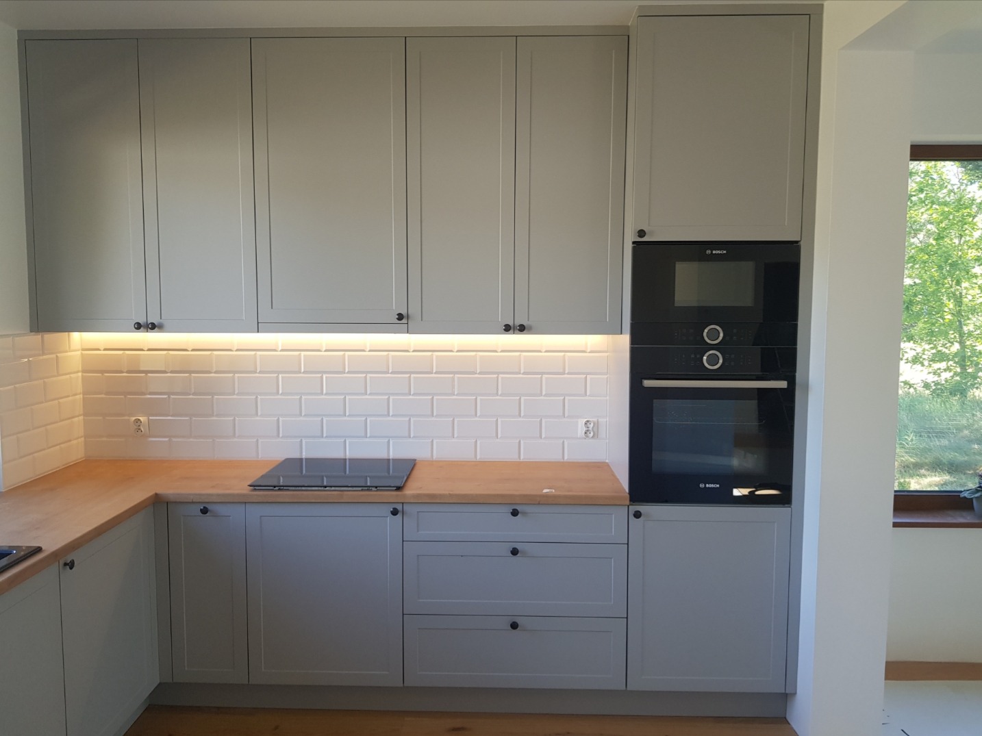Szara zabudowa kuchenna z drewnianym blatem, białymi kafelkami typu metro, wbudowanym piekarnikiem i płytą indukcyjną, oświetleniem podszafkowym LED oraz widokiem na zieleń za oknem.
