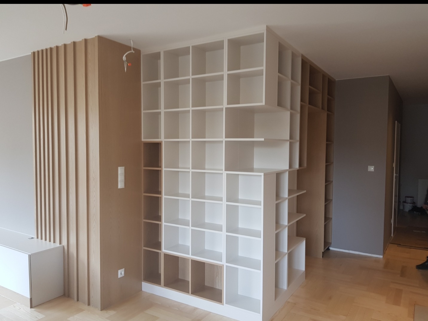 Wielopoziomowa, biała szafa wnękowa z otwartymi półkami, częściowo zabudowana, z drewnianymi elementami dekoracyjnymi na ścianie i parkietem na podłodze, widoczne przewody elektryczne.