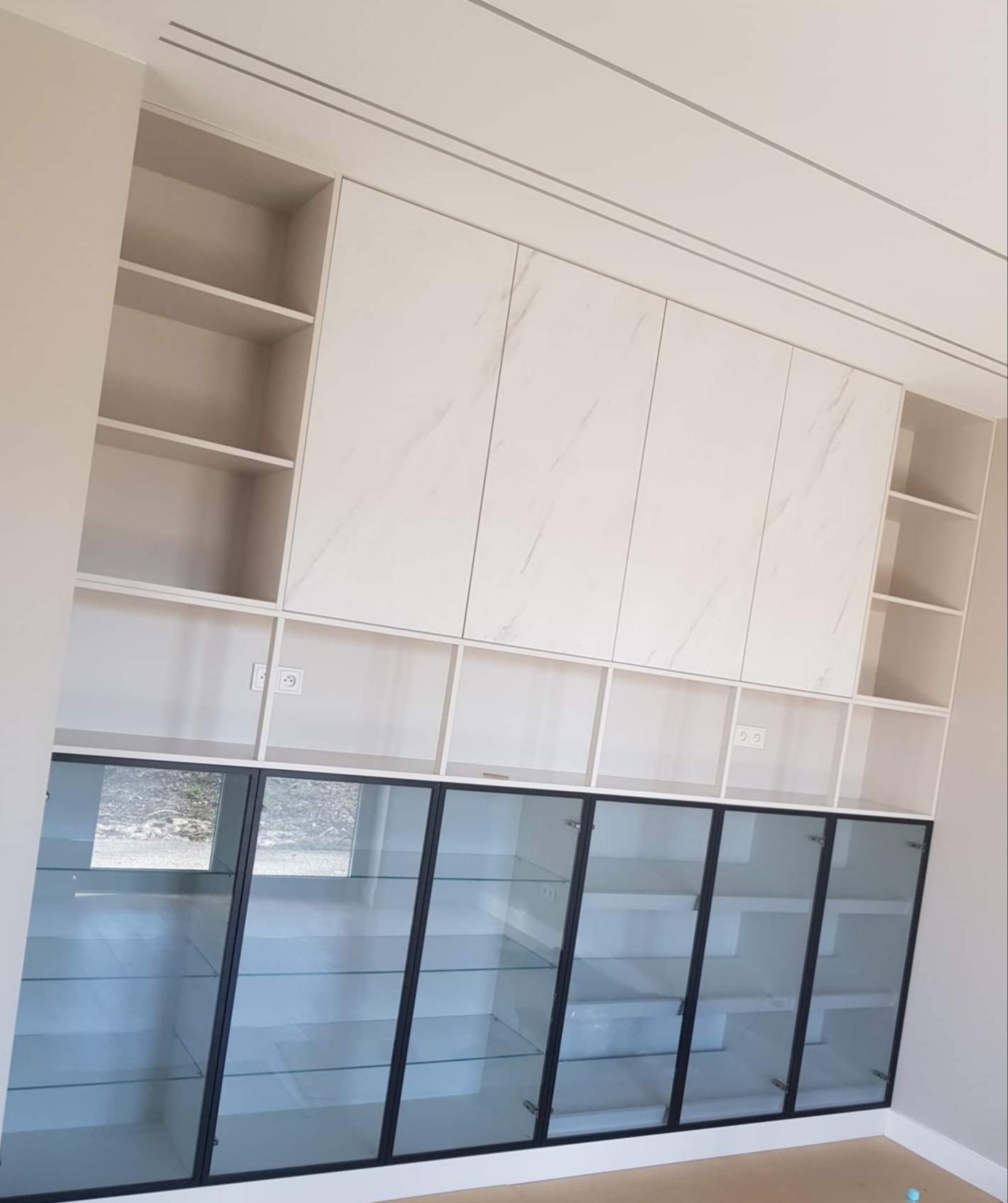 Biała zabudowa meblowa z przeszklonymi drzwiczkami i marmurkowymi frontami, widoczne gniazdka elektryczne w środkowej części, ujęcie z dołu.