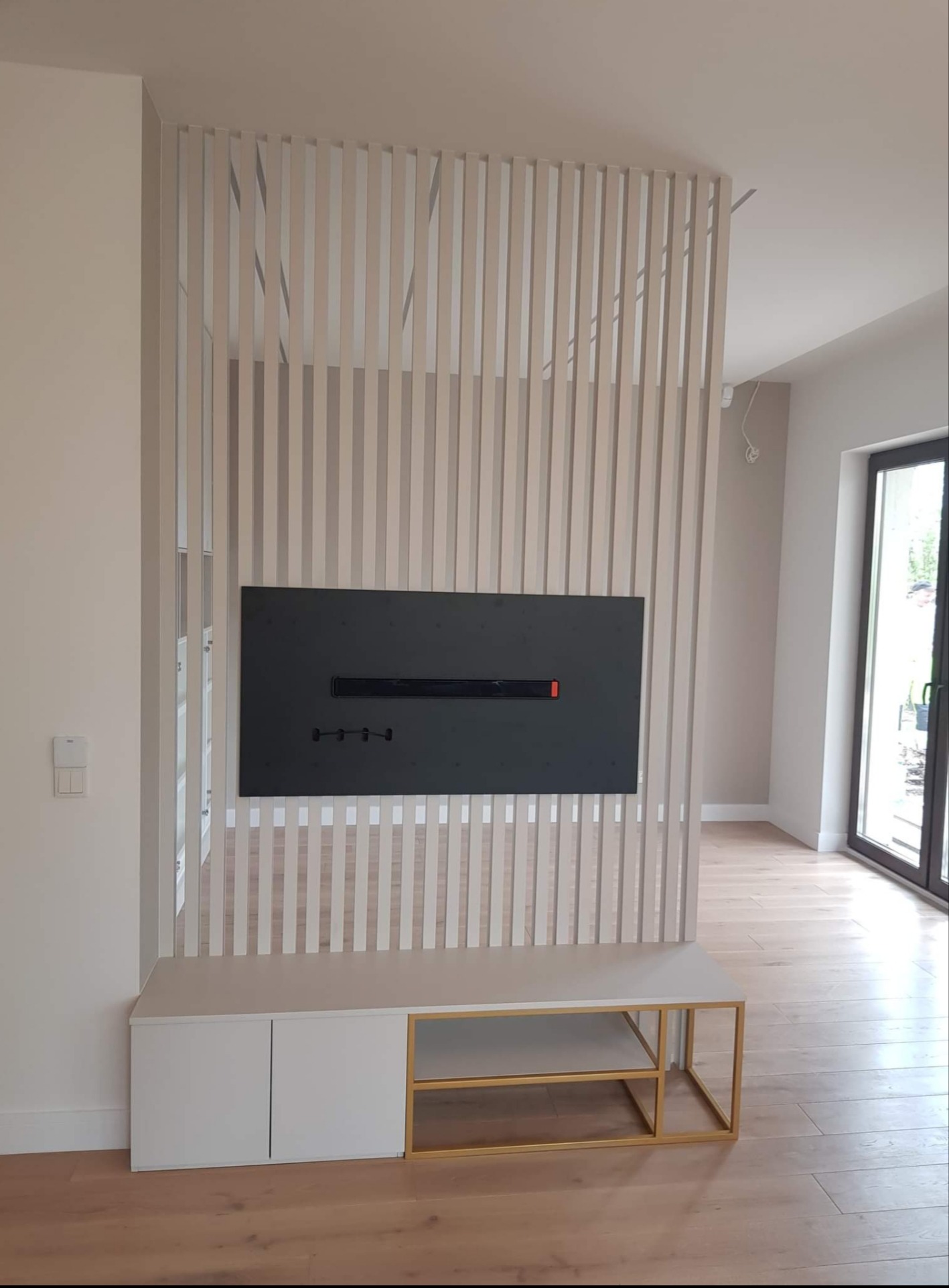 Nowoczesny salon z drewnianą podłogą i ażurową ścianką działową z pionowych listew w kolorze beżowym, za którą widoczny jest korytarz. Na ściance zawieszony czarny panel z wbudowanym biokominkiem.