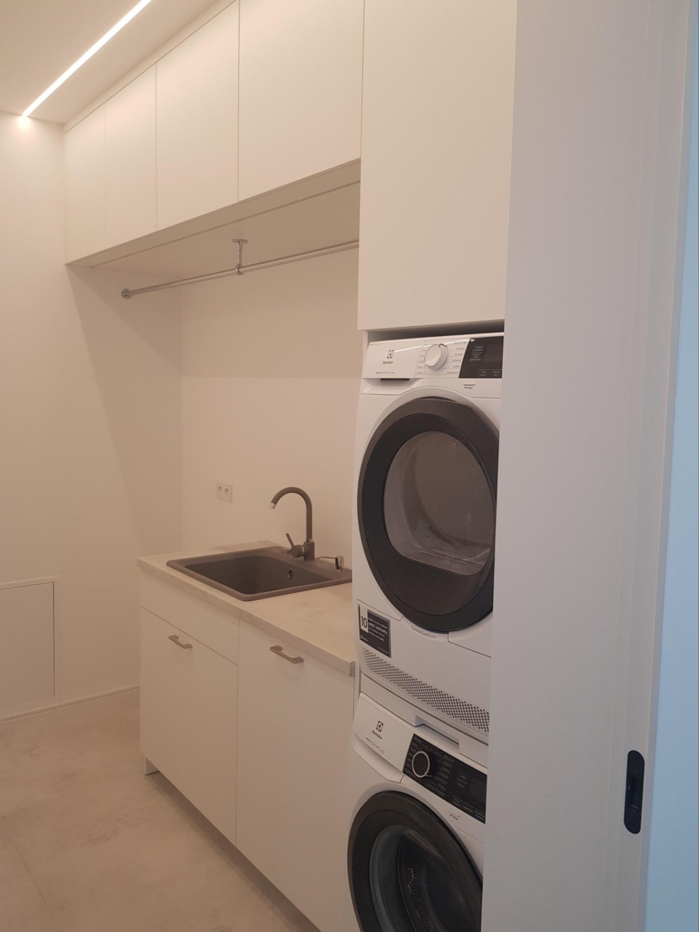 Nowoczesna pralnia z białymi szafkami, ciemnoszarym zlewem i pralko-suszarką Electrolux w zabudowie, oświetlenie LED podszafkowe, minimalistyczny design.