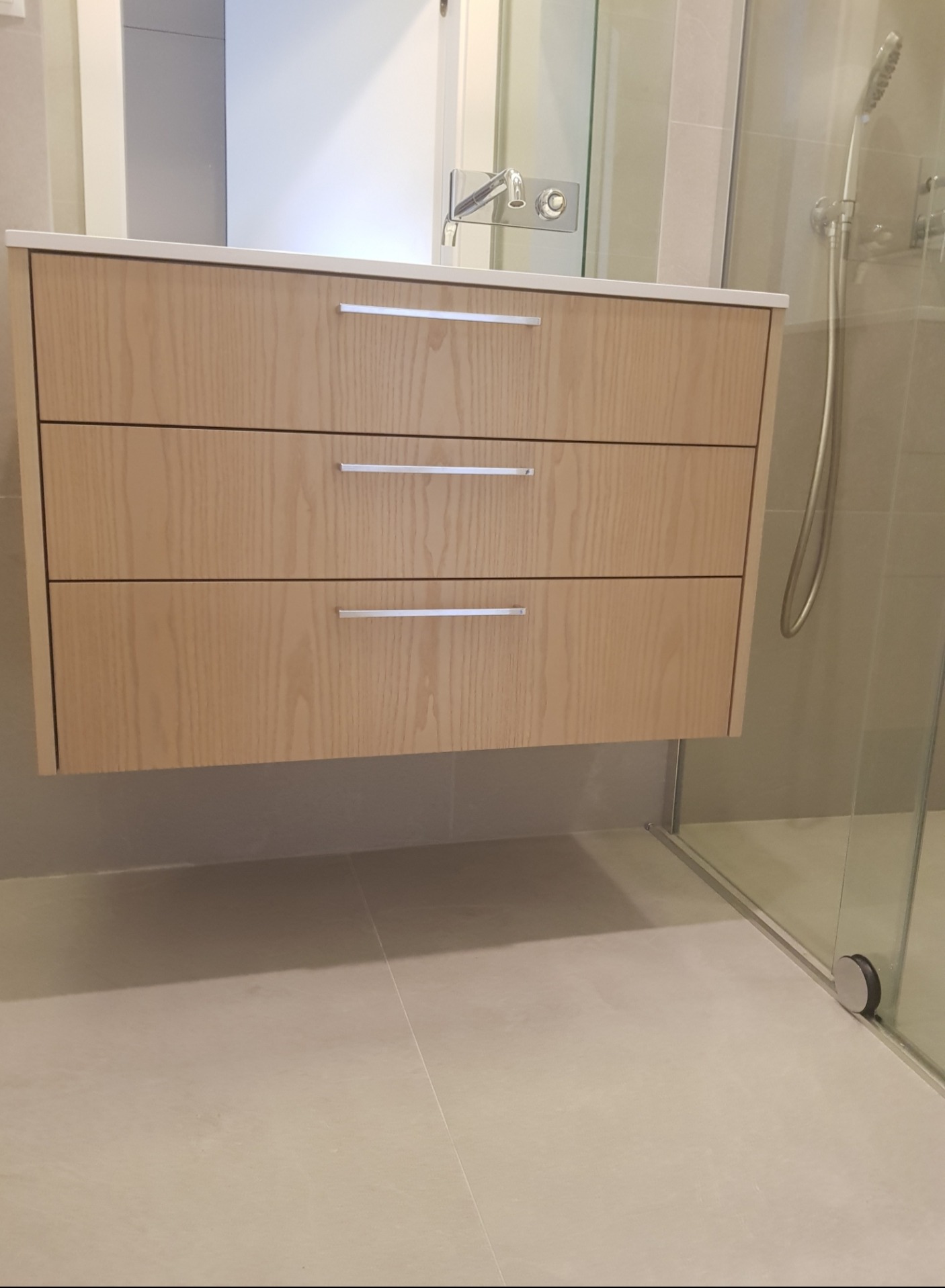 Nowoczesna, podwieszana szafka łazienkowa z trzema szufladami o jasnym, drewnianym wykończeniu, z chromowanymi uchwytami, zamontowana pod umywalką w minimalistycznej łazience z kabiną prysznicową...