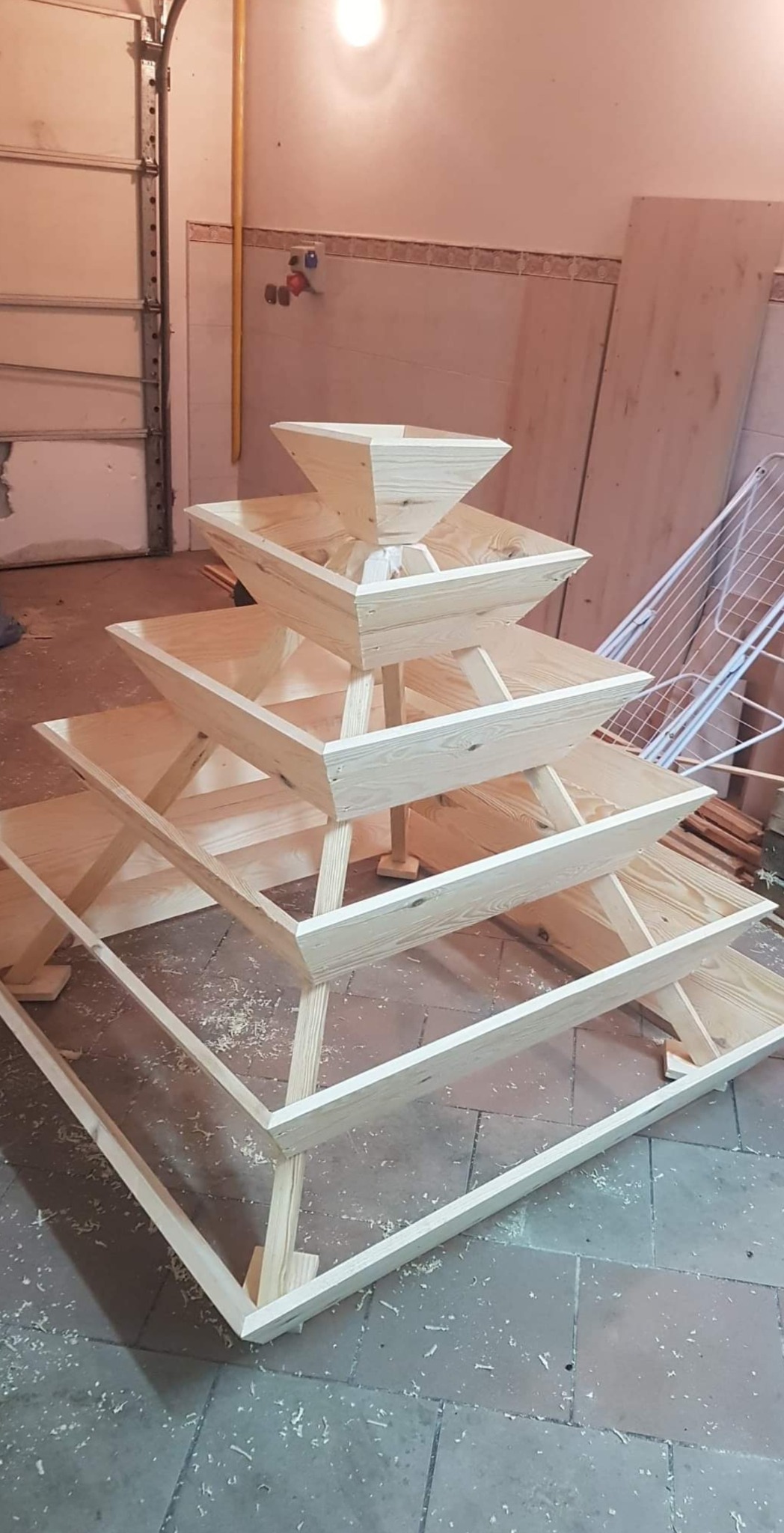 Surowa, drewniana konstrukcja schodkowa z kwadratowymi pojemnikami, prawdopodobnie do ekspozycji, w warsztacie z płytkami podłogowymi i garażową bramą w tle, pokryta wiórami.