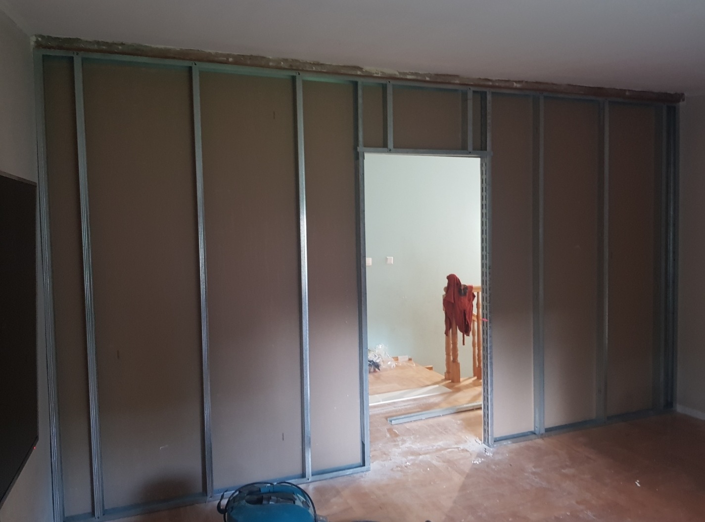 Konstrukcja ścianki działowej z metalowych profili i płyt kartonowo-gipsowych w trakcie montażu, widoczne narzędzie elektryczne na podłodze, otwór drzwiowy w przygotowanej konstrukcji.