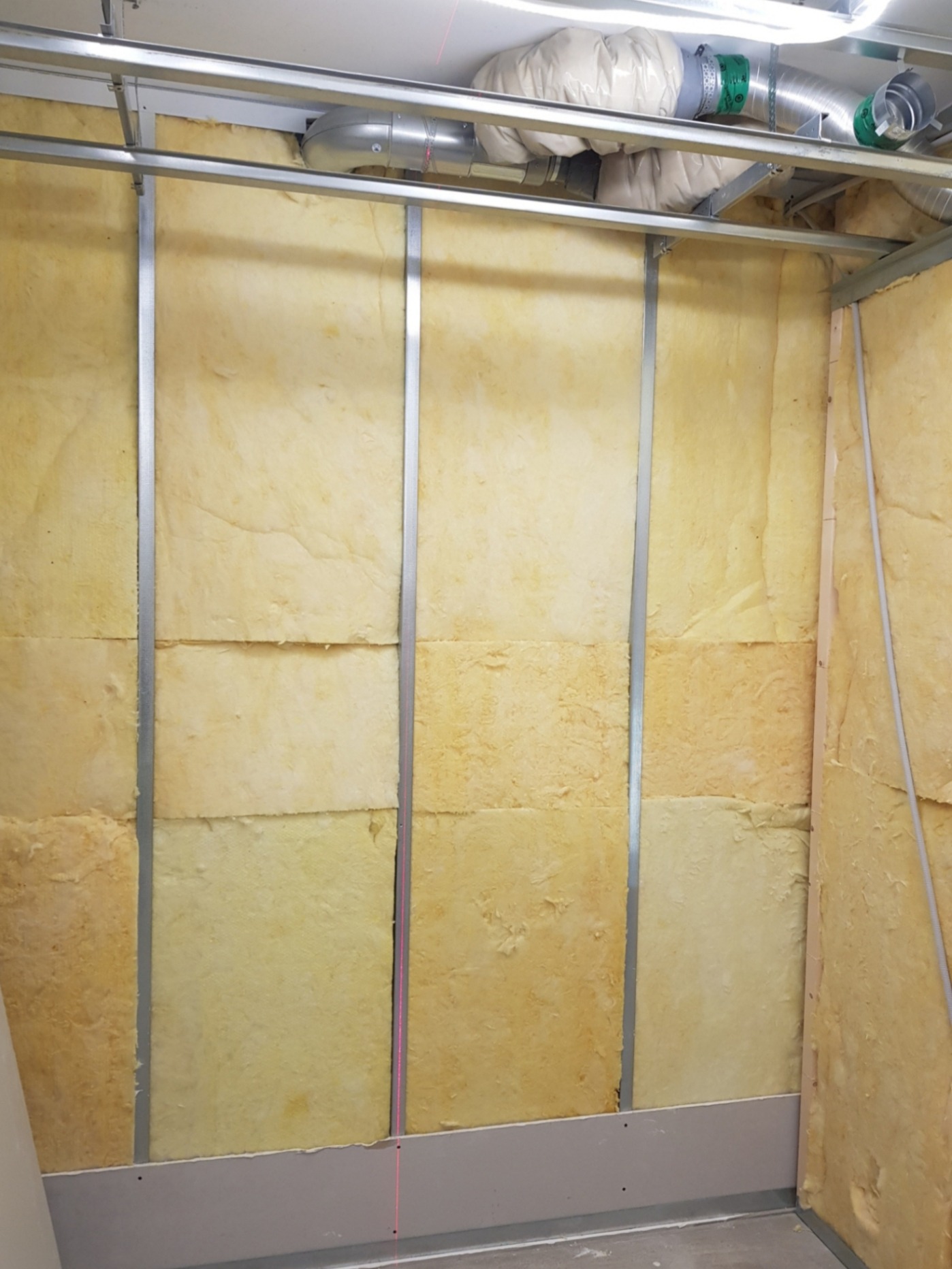 Ściana w trakcie izolacji wełną mineralną, widoczne metalowe profile konstrukcyjne, sufit z instalacją wentylacyjną i fragment lasera krzyżowego.