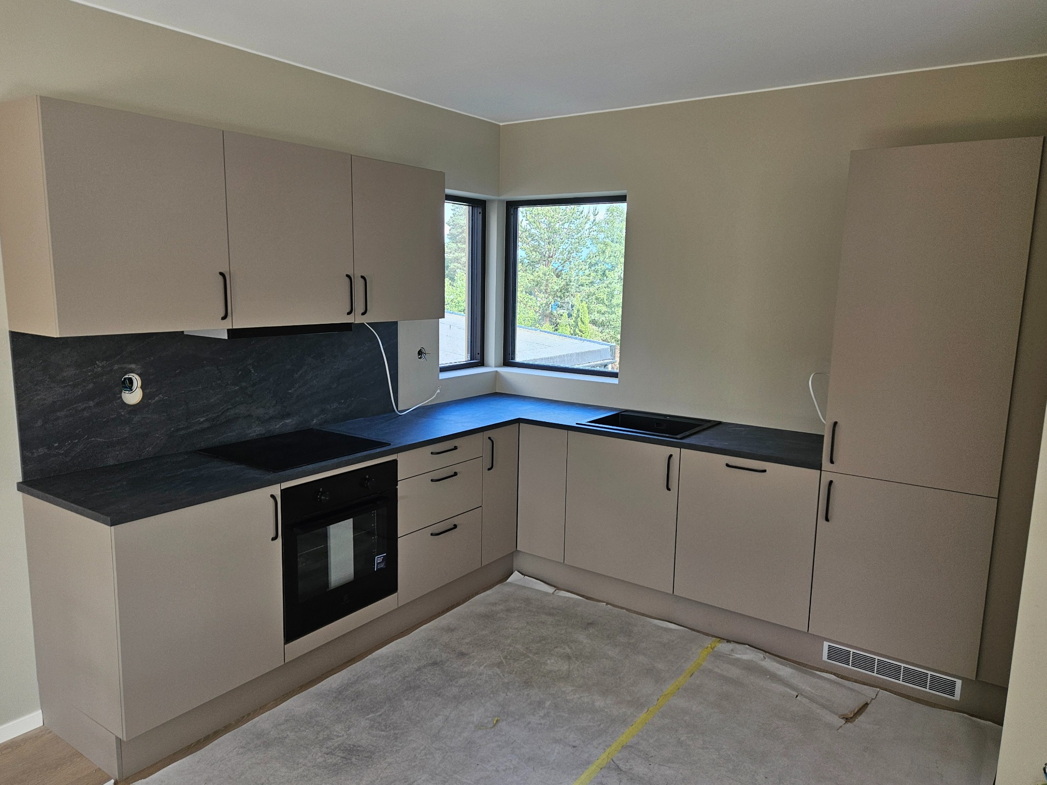 Nowoczesna kuchnia z beżowymi frontami szafek i czarnym blatem, widok na okno z zielenią na zewnątrz, podłoga zabezpieczona folią malarską.