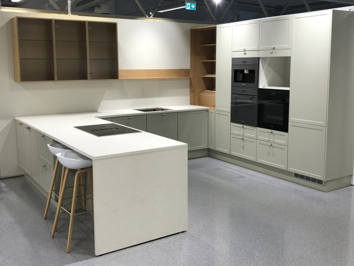 Jasnoszara zabudowa kuchenna z wyspą, białym blatem i dwoma białymi hokerami na drewnianych nogach, ekspozycja w salonie meblowym.