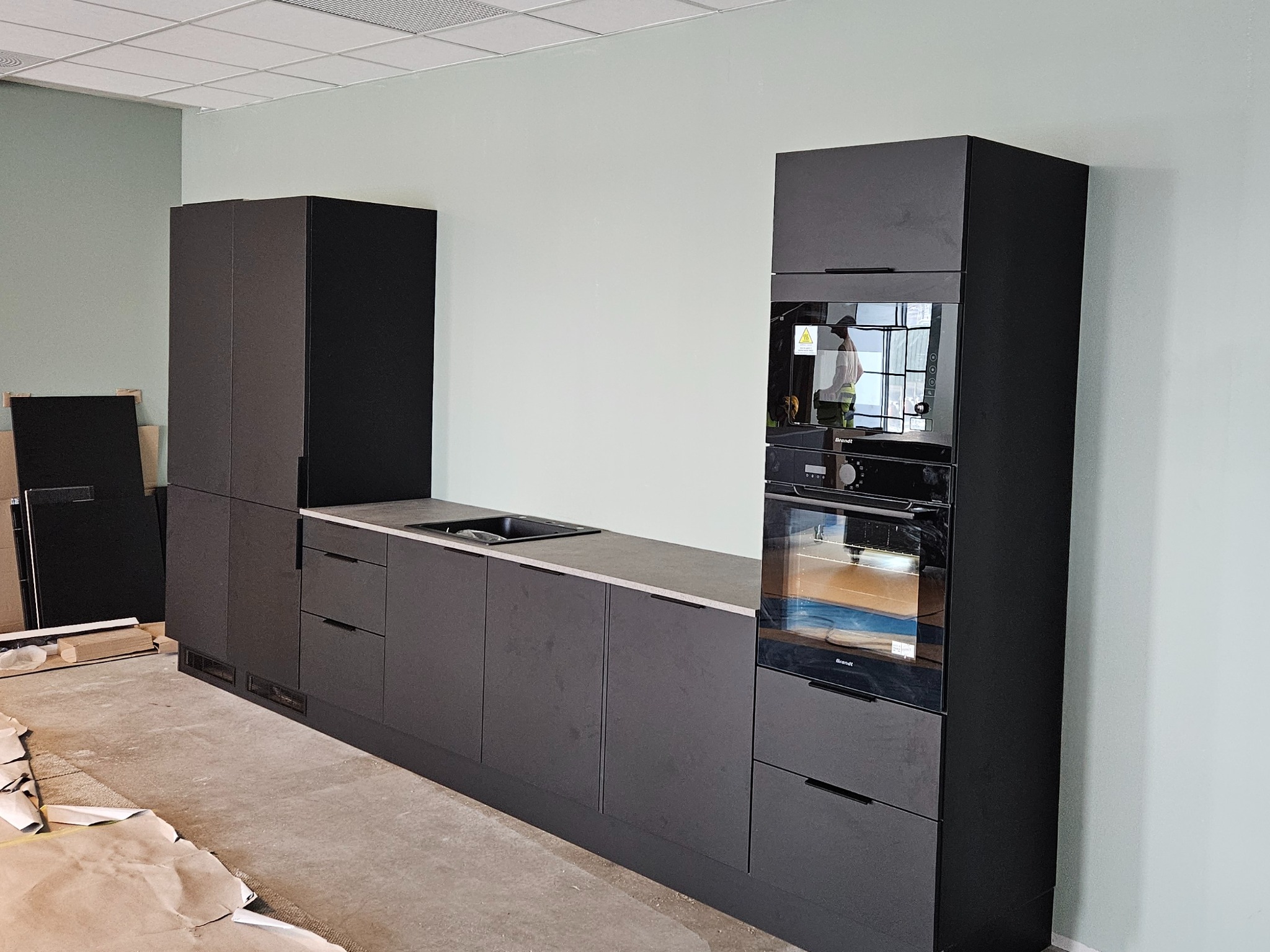 Ciemnoszara zabudowa kuchenna z piekarnikiem i szafkami, widoczne zabezpieczenia podłogi podczas montażu, surowy stan wykończenia wnętrza.