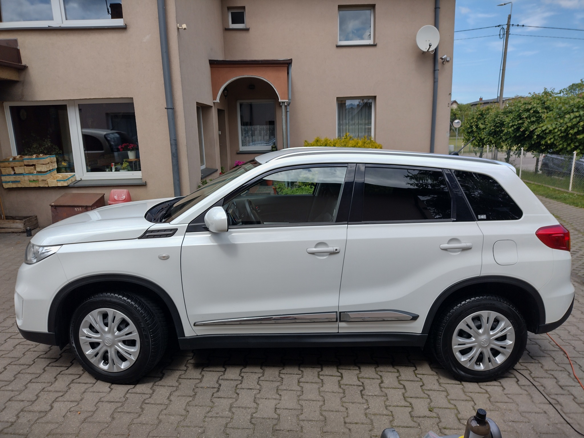 Biały samochód typu SUV marki Suzuki zaparkowany na brukowanym podjeździe przed beżowym domem z widoczną anteną satelitarną.