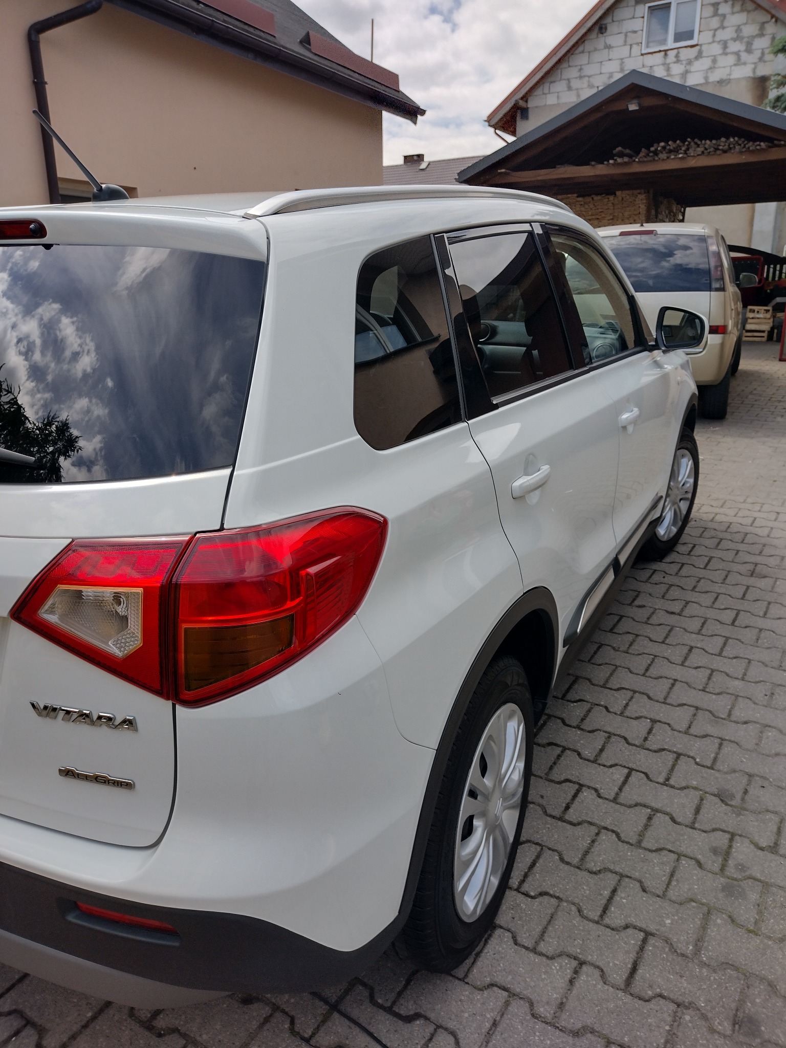 Biała Suzuki Vitara zaparkowana na kostce brukowej, widok z tyłu, z odbiciem chmur w szybie. Drugi pojazd widoczny w tle.