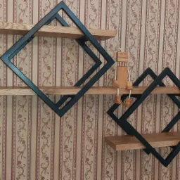 Dwie industrialne półki ścienne w kształcie rombów z czarną metalową ramą i drewnianymi blatami na tle tapety w paski z motywem kwiatowym, z małą drewnianą figurką siedzącą na dolnej półce.