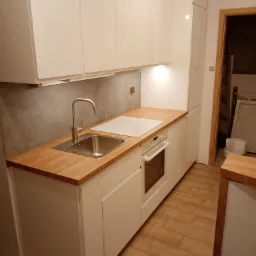 Nowoczesna, jasna kuchnia z białymi, błyszczącymi frontami szafek, drewnianym blatem, stalowym zlewem i płytą indukcyjną. Widoczny fragment podłogi z paneli imitujących drewno oraz oświetlenie...