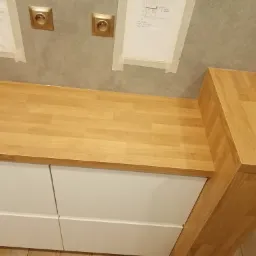 Fragment blatu z jasnego drewna na białych szafkach z zamontowanymi gniazdkami elektrycznymi w tle i planami na ścianie.
