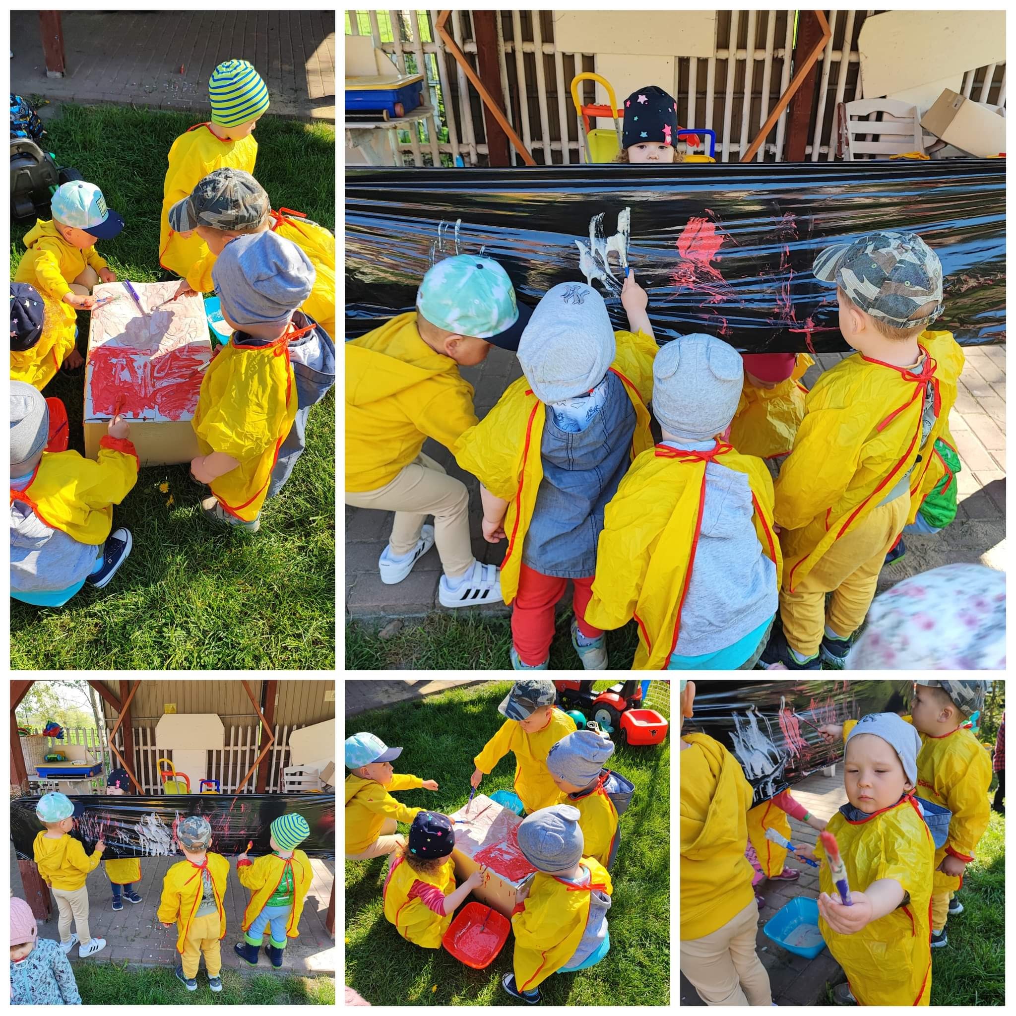 Grupa dzieci w żółtych fartuchach maluje farbami na kartonie i czarnej folii na zewnątrz, pod nadzorem opiekuna, w tle widoczne zabawki.