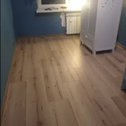 Beżowe panele podłogowe ułożone w pokoju z niebieskimi ścianami, widoczny fragment białej szafy i grzejnika pod oknem.